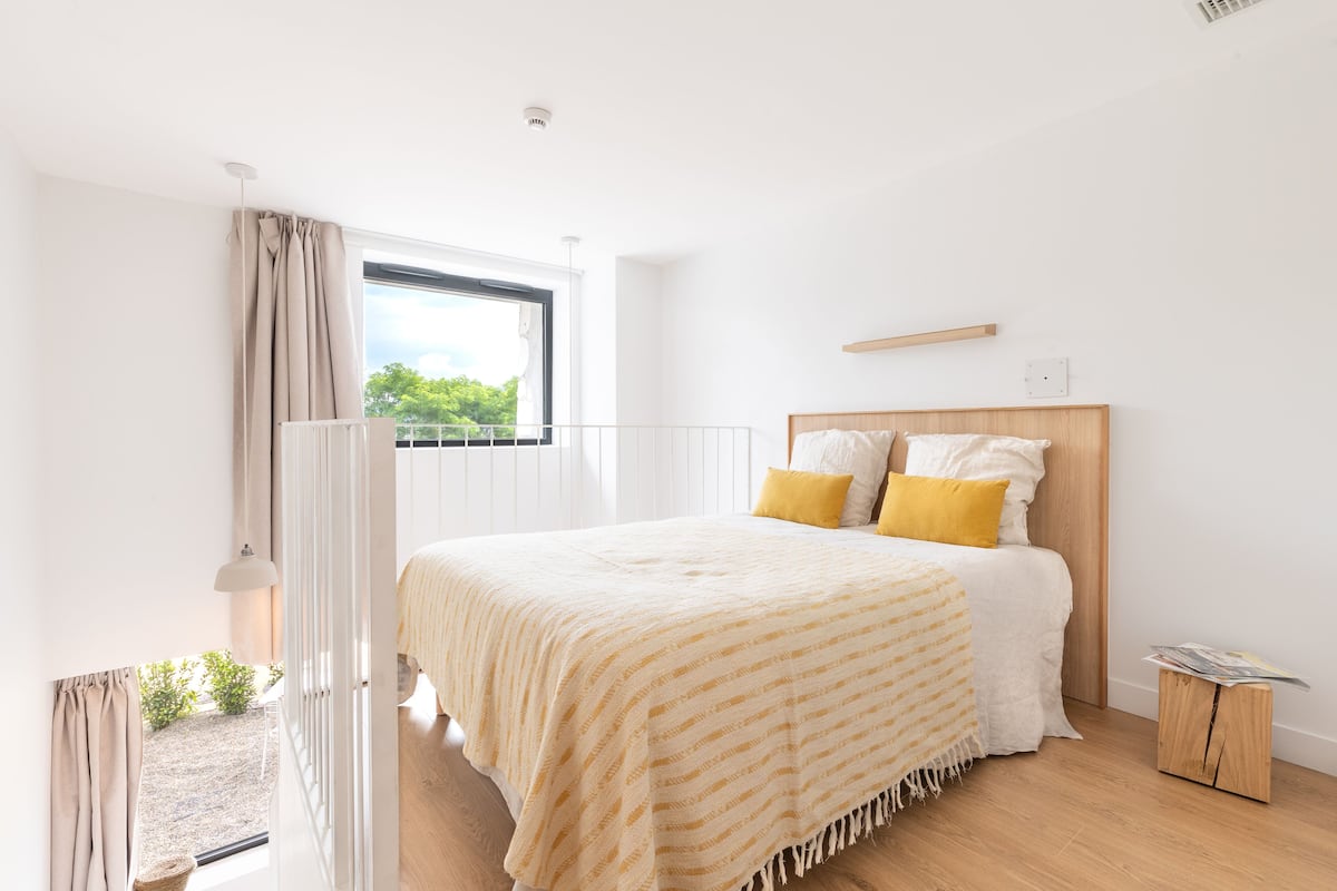 A bedroom with a large bed, a window with curtains, a bedside table, and a view of greenery outside.