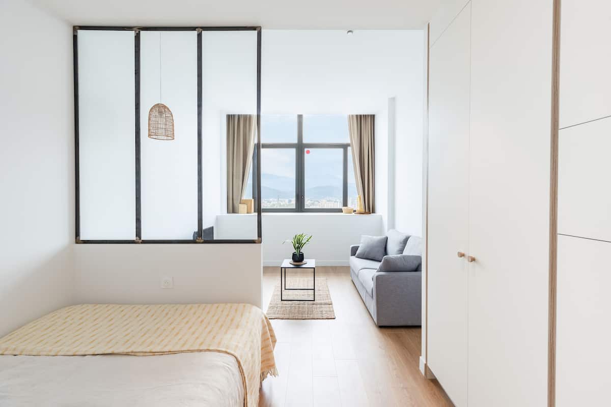 A modern, minimalist room with a bed, a sofa, and a large window offering a city view.