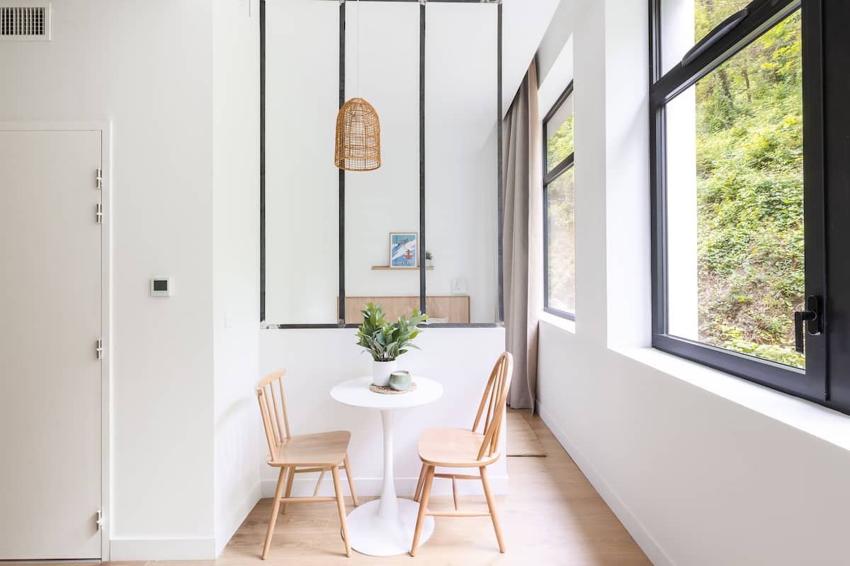 A modern room with a white door, a round table with two chairs, a hanging wicker light, and a view of greenery through large windows.