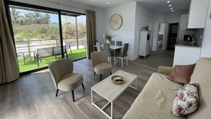 Ett modernt vardagsrum med en soffa, fåtöljer, ett soffbord och en matplats med ett bord och stolar. Det finns också ett köksområde med skåp och ett kylskåp. Rummet har stora fönster med utsikt över en trädgård och en balkong.
