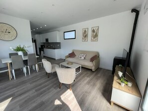 Ein modernes Wohnzimmer mit einer Couch, einem Couchtisch und Sesseln. Es gibt einen Essbereich mit Tisch und Stühlen. Im Hintergrund ist eine Küche zu sehen.