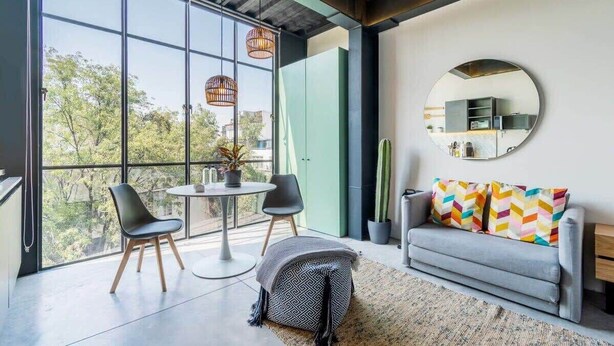 A modern living room with large windows, a round mirror, a grey sofa with colorful cushions, and a small round table with two chairs. - w* | Glorious Loft w/ Beautiful Deco in Roma Norte (Ciudad de México)