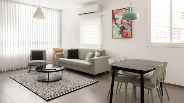 A modern living room with a grey sofa, a round coffee table, and a dining table with chairs. There is a colorful artwork on the wall and an air conditioning unit above the sofa. - Amazing APT Bat-Yam Near Beach (Bat Yam)