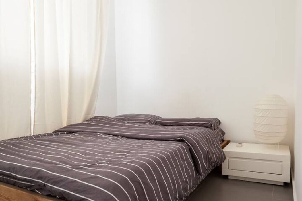 A neatly made bed with striped bedding, a white bedside table with a lamp, and a window with sheer curtains.