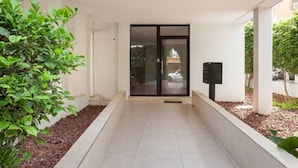 A modern building entrance with a tiled pathway, a glass door, and a small garden area with a tree. - Amazing APT Bat-Yam Near Beach (Bat Yam)