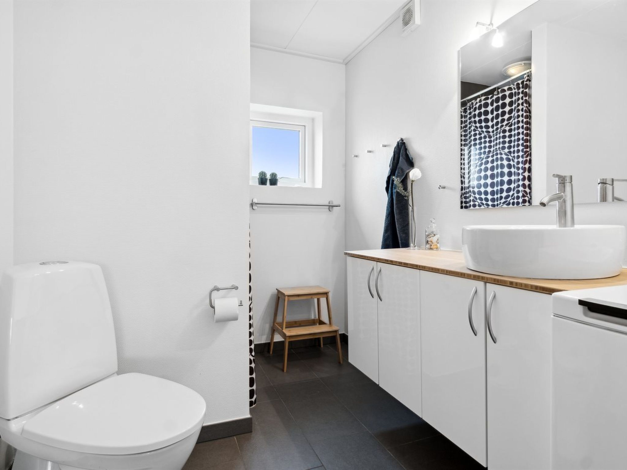 Een moderne badkamer met een wit toilet, een houten krukje, een ruime wastafel met spiegel en een raam met uitzicht op de lucht.
