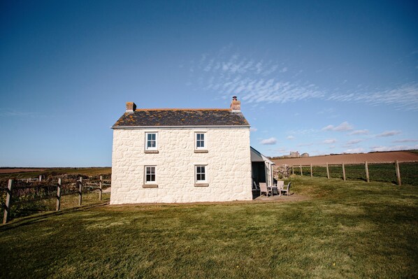 A white stone house with a sloped roof and a chimney, set in a grassy field with a clear sky. - Faraway Cottage (Penzance)