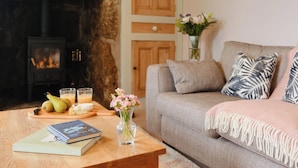 A cozy living room with a fireplace, a sofa, a coffee table with books and a vase of flowers, and a wooden sideboard with fruits and drinks. - Bosistow Farmhouse (Penzance)