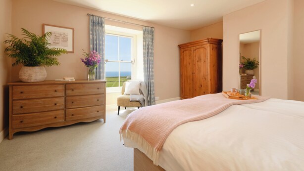 A bedroom with a large bed, a wooden dresser, a chair, a large mirror, and a view of the outdoors. - Bosistow Farmhouse (Penzance)