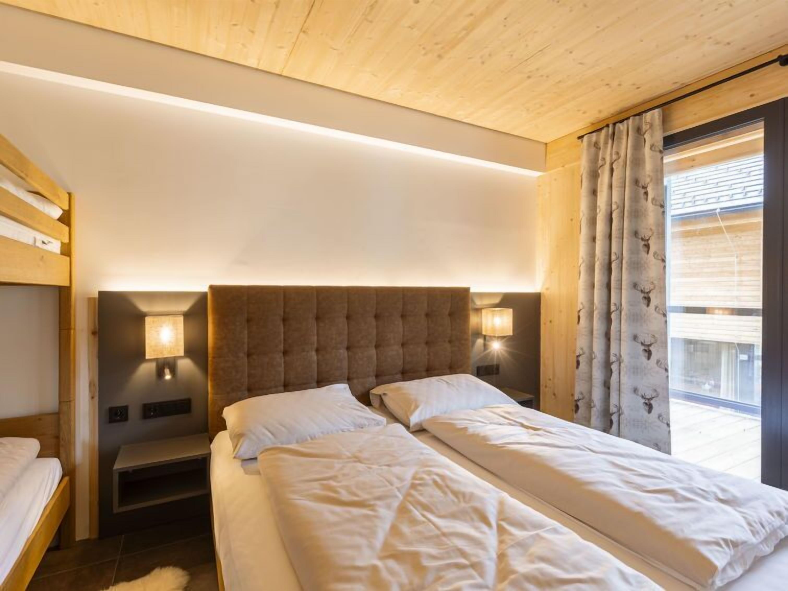 Habitación de hotel con techo de madera, dos camas con ropa blanca, una mesita de noche con lámpara y una ventana con cortinas.