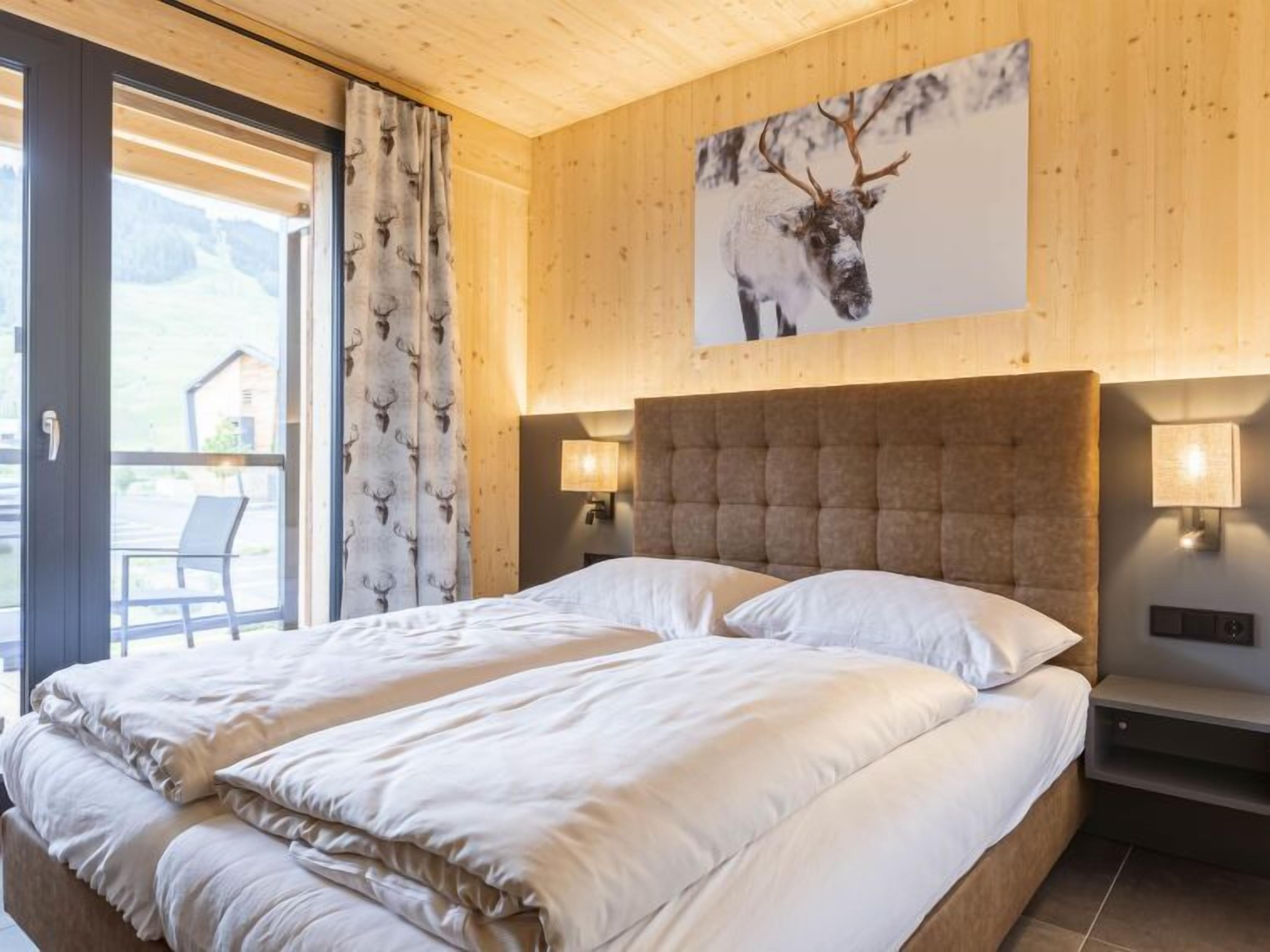 Un lit double avec une tête de lit, deux lampes de chevet, une fenêtre avec des rideaux et un tableau représentant un cerf accroché au mur.