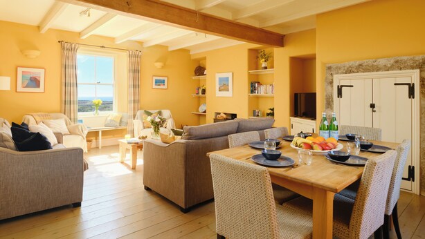 A spacious living area with a sofa, dining table, and chairs. There is a window with curtains, a bookshelf, and a door with a coat rack. - Higher Roskestal (Penzance)