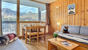 A cozy room with a wooden interior, a sofa, a dining table with chairs, and a large window offering a mountain view.