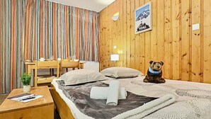 A hotel room with a wooden paneled wall, a bed with a white comforter and pillows, a small desk with a chair, a table with a lamp, and a framed picture on the wall.