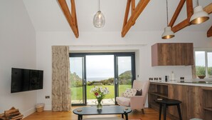 A living room with a TV, a dining area with a table and chairs, a kitchen with wooden cabinets, and a large sliding glass door leading to a garden. - Pendower Cottage (Penzance)