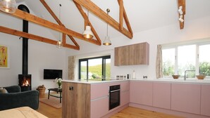 A modern kitchen with wooden beams, pink cabinets, and a large window. - Pendower Cottage (Penzance)