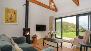 A living room with a fireplace, a sofa, a coffee table, and a view of the outdoors. - Pendower Cottage (Penzance)