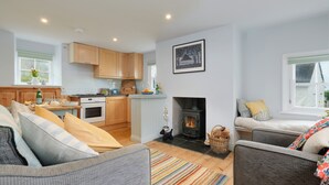 A modern kitchen with wooden cabinets, a dining area with a white table, and a cozy seating area with a fireplace. - Corner Cottage (Penzance)