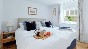 A neatly made bed with a tray of breakfast items, a bedside table with a lamp, and a window with curtains. - Corner Cottage (Penzance)