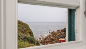 A coastal view through a window, featuring a rocky shoreline, a grassy cliff, and the sea. - Corner Cottage (Penzance)