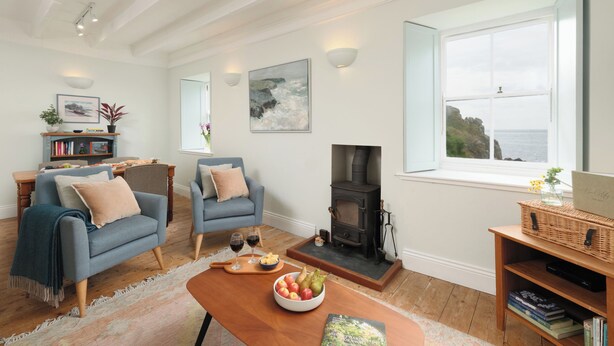 A cozy living room with a fireplace, two blue armchairs, a wooden coffee table with fruit, and a bookshelf. - Cove Cottage (Penzance)