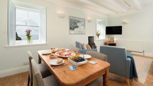 A dining area with a wooden table set for a meal, a sofa, and a TV in the background. - Cove Cottage (Penzance)