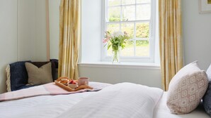 A neatly made bed with a tray of breakfast items, a vase of flowers by the window, and a view of greenery outside. - Cove Cottage (Penzance)