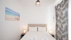A neatly made bed with white linens and rolled towels, a painting of a beach scene, and a patterned curtain. - Island Dreaming Opal 2 Bd w shared pool (Mykonos)