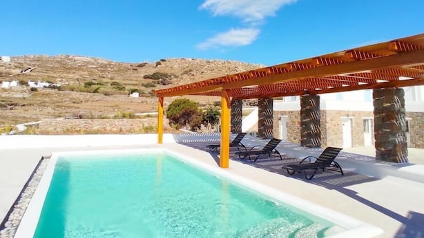 A rectangular swimming pool with clear blue water, surrounded by a white deck. There are lounge chairs under a wooden pergola. The background features a hilly landscape with a clear sky. - Island Dreaming Opal 2 Bd w shared pool (Mykonos)