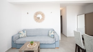 A modern living room with a light blue sofa, two patterned cushions, a wooden coffee table with a plant, and a decorative mirror on the wall. - Island Dreaming Opal 2 Bd w shared pool (Mykonos)