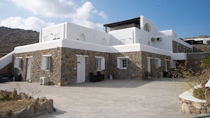 A modern building with a stone facade and white upper levels, featuring a spacious courtyard with outdoor seating and a clear view of the surrounding landscape. - Island Dreaming Opal 2 Bd w shared pool (Mykonos)