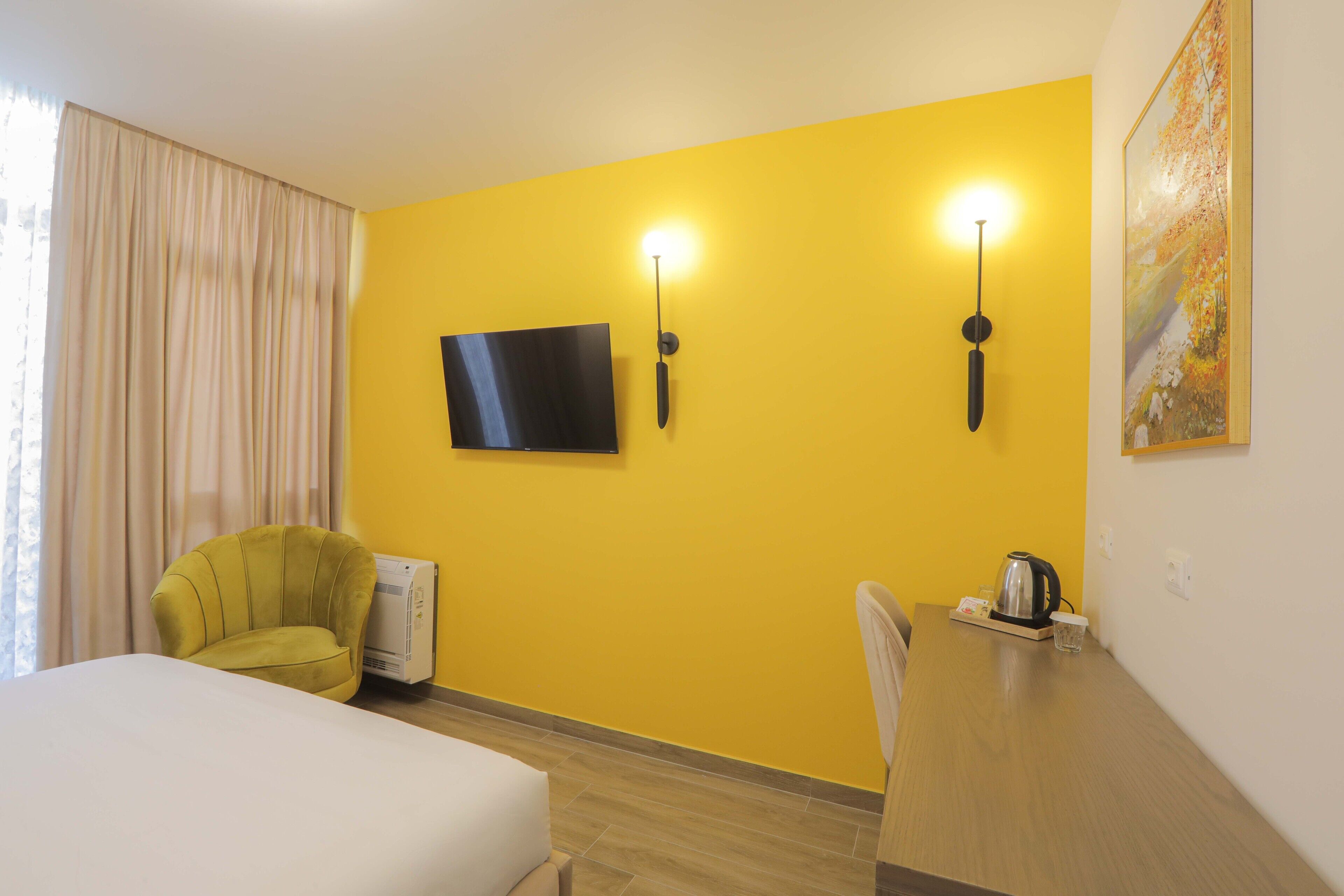A hotel room with a yellow wall, a flat-screen TV, a bed, a desk with a kettle, and a chair.