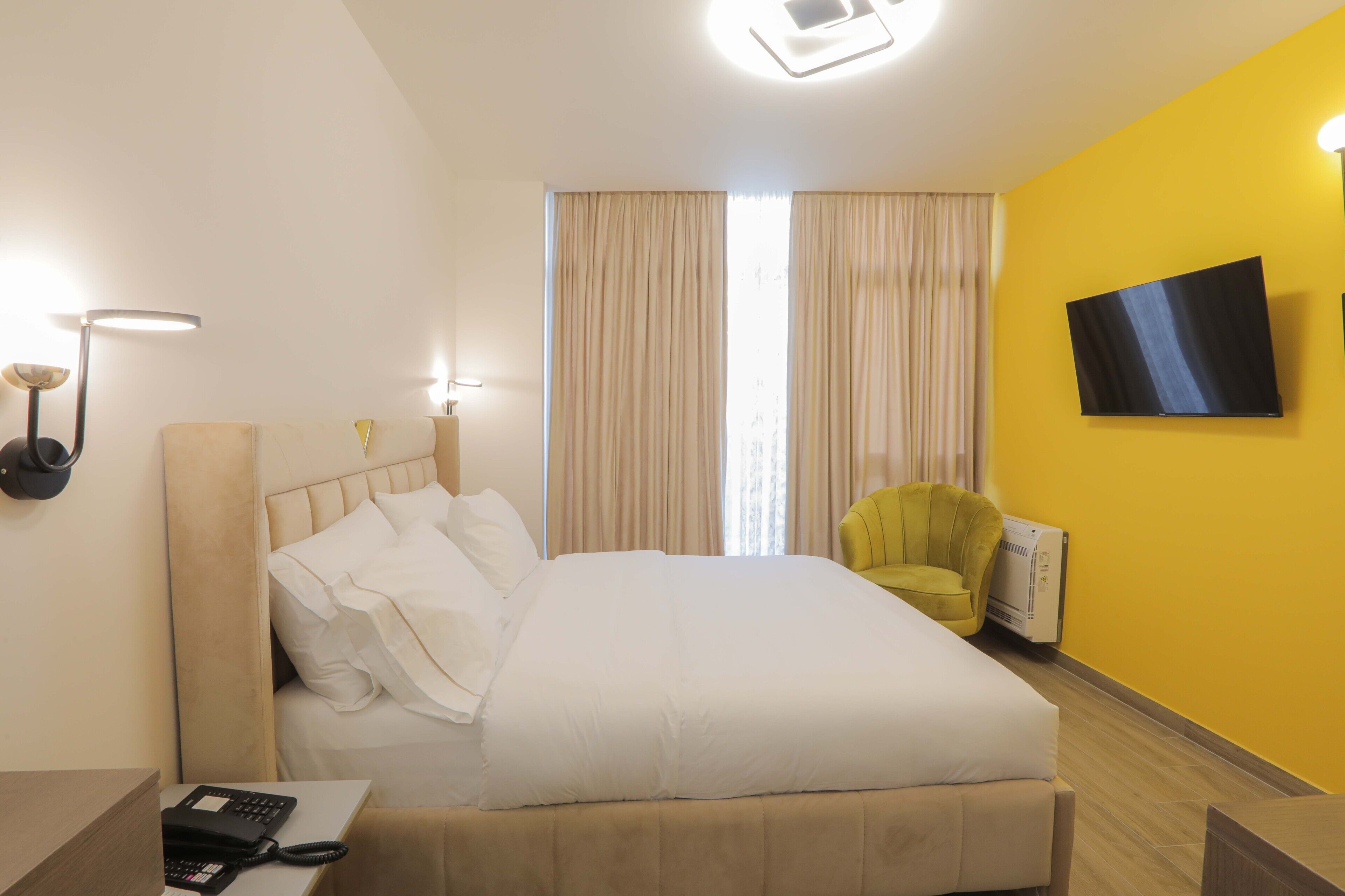 A hotel room with a bed, a desk with a telephone, a chair, a television on the wall, and curtains.