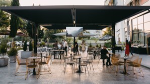 An outdoor cafe with multiple tables and chairs, surrounded by potted plants and a covered seating area. - Olia Residence (Tirana)