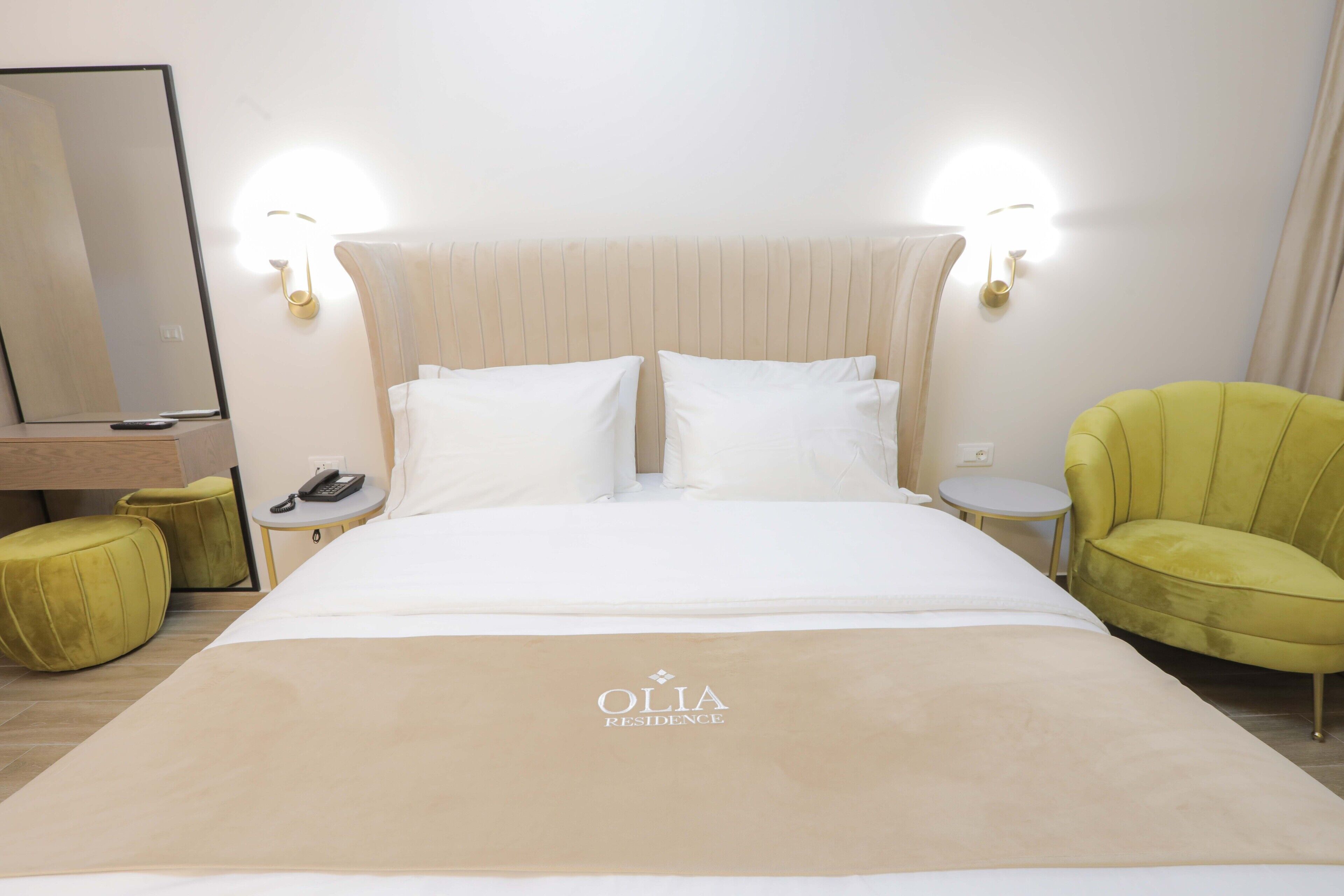 Habitación de hotel con una cama, dos mesitas de noche, un espejo, un sillón verde y una lámpara fijada a la pared.