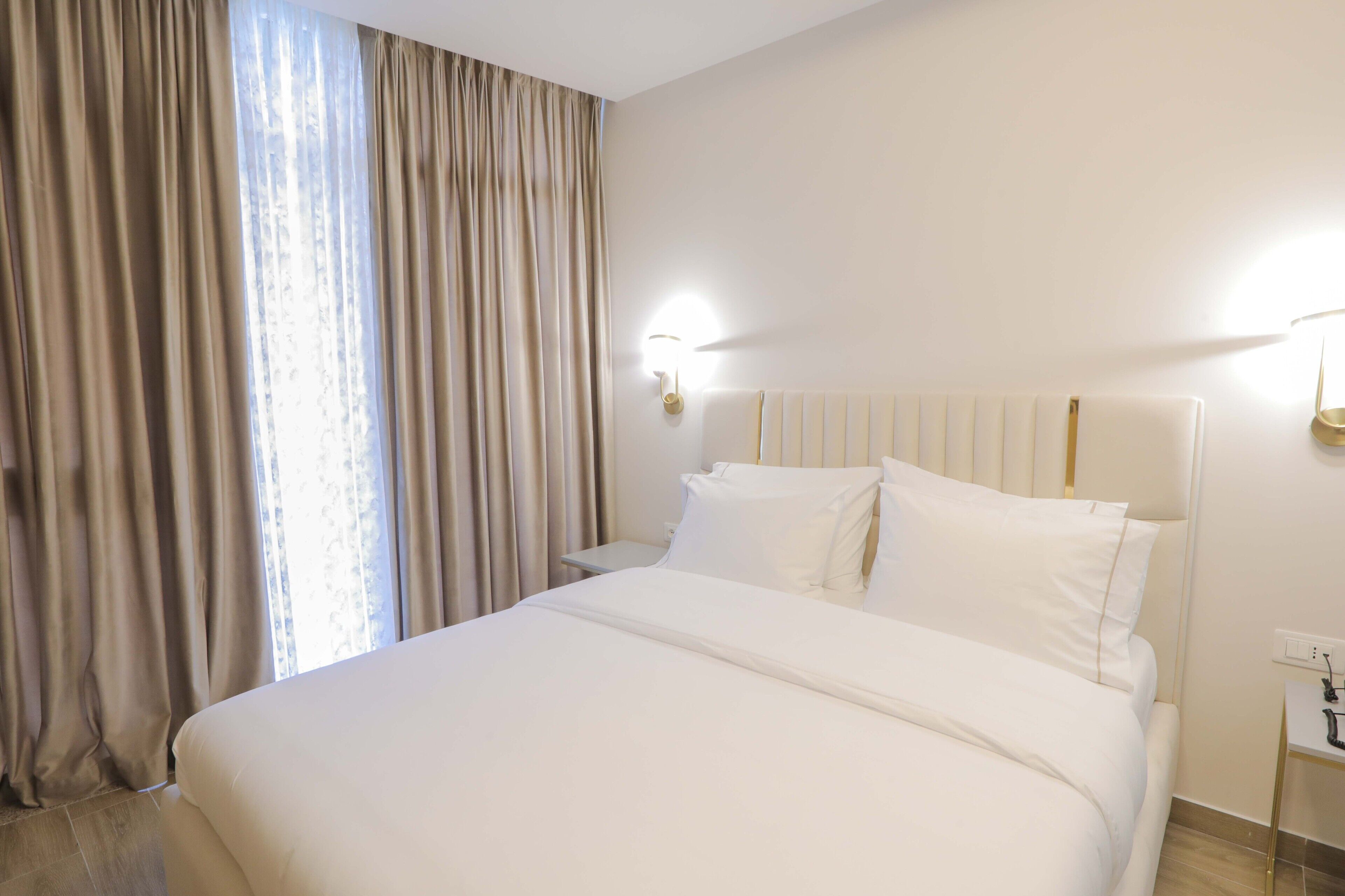 Quarto de hotel com uma cama grande, mesa de cabeceira, abajur e janela com cortinas.