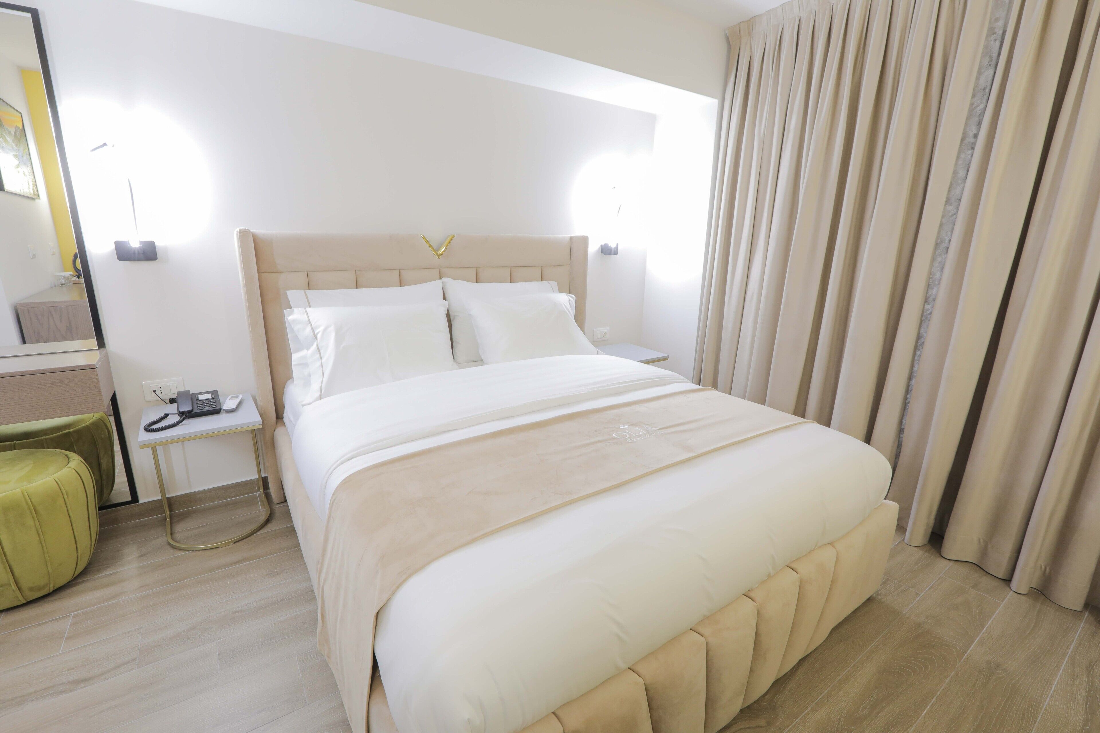 A hotel room with a large bed, a bedside table with a phone, a green ottoman, and a mirror.
