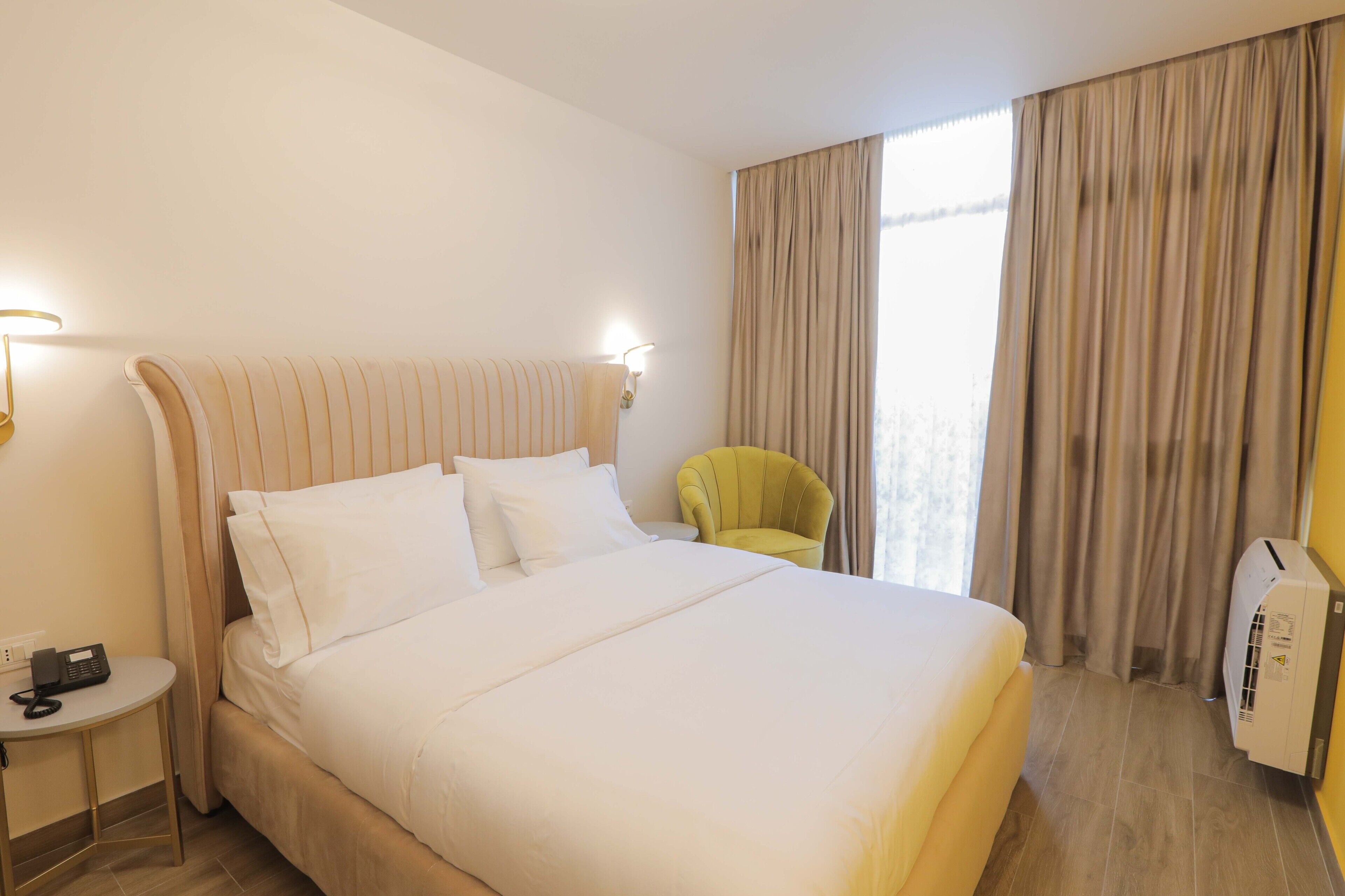 A hotel room with a large bed, a bedside table with a phone, a yellow armchair, and a window with curtains.