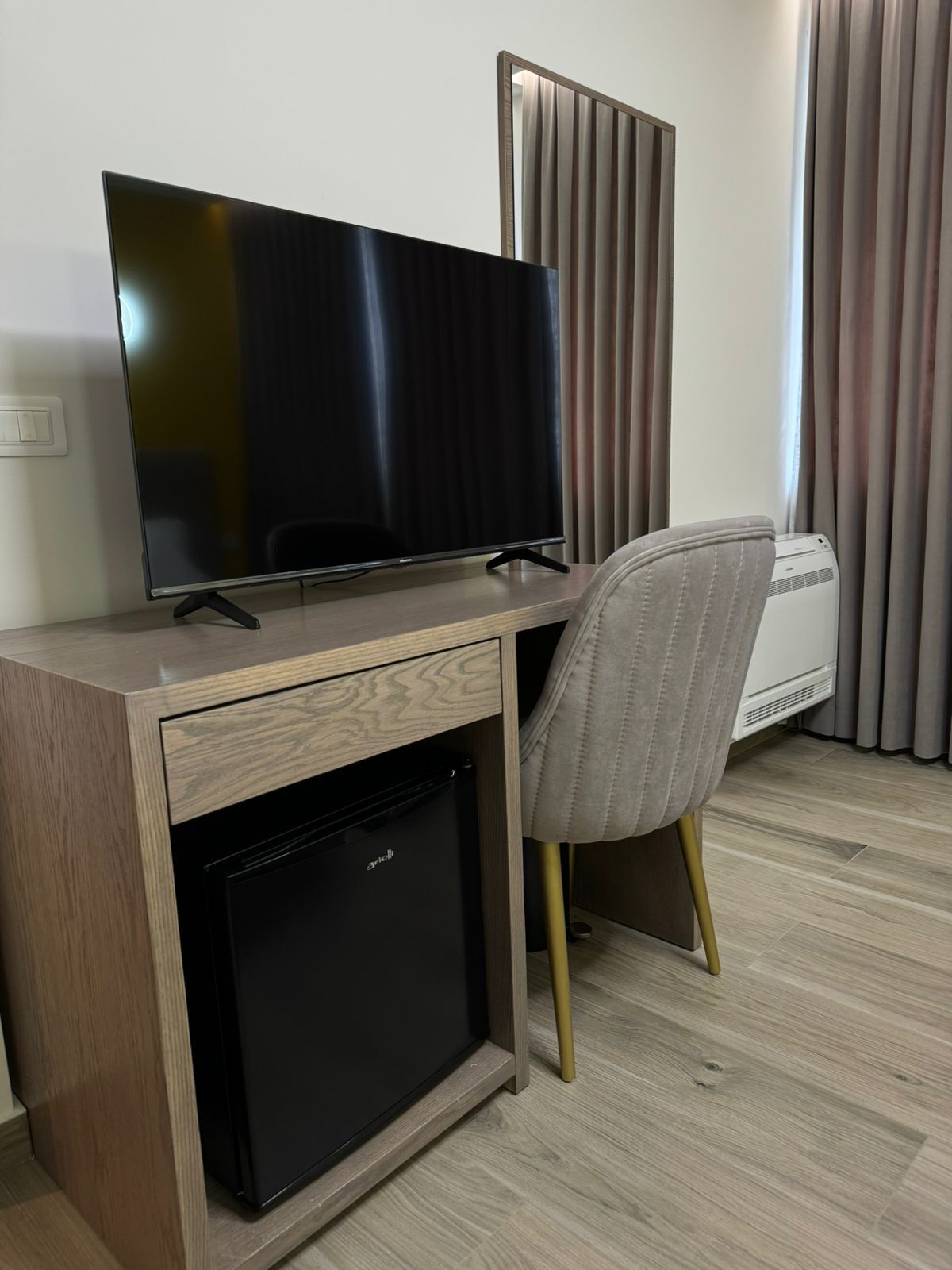 Ett modernt hotellrum med ett skrivbord i trä, en platt-TV, ett litet kylskåp, en grå kontorsstol och en vit luftkonditioneringsanläggning.