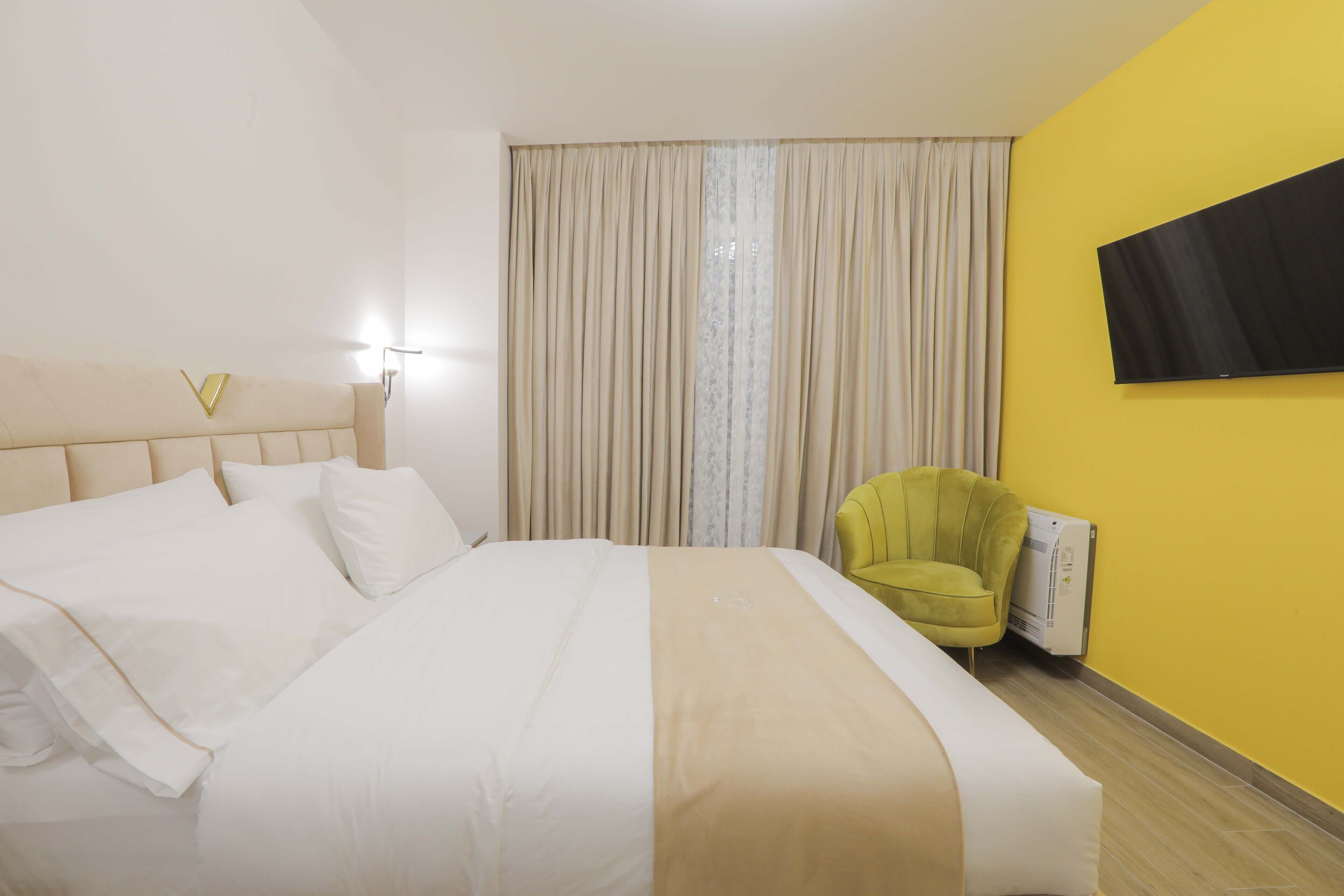 Ett hotellrum med en stor säng, en gul vägg, en platt-TV och en grön fåtölj.