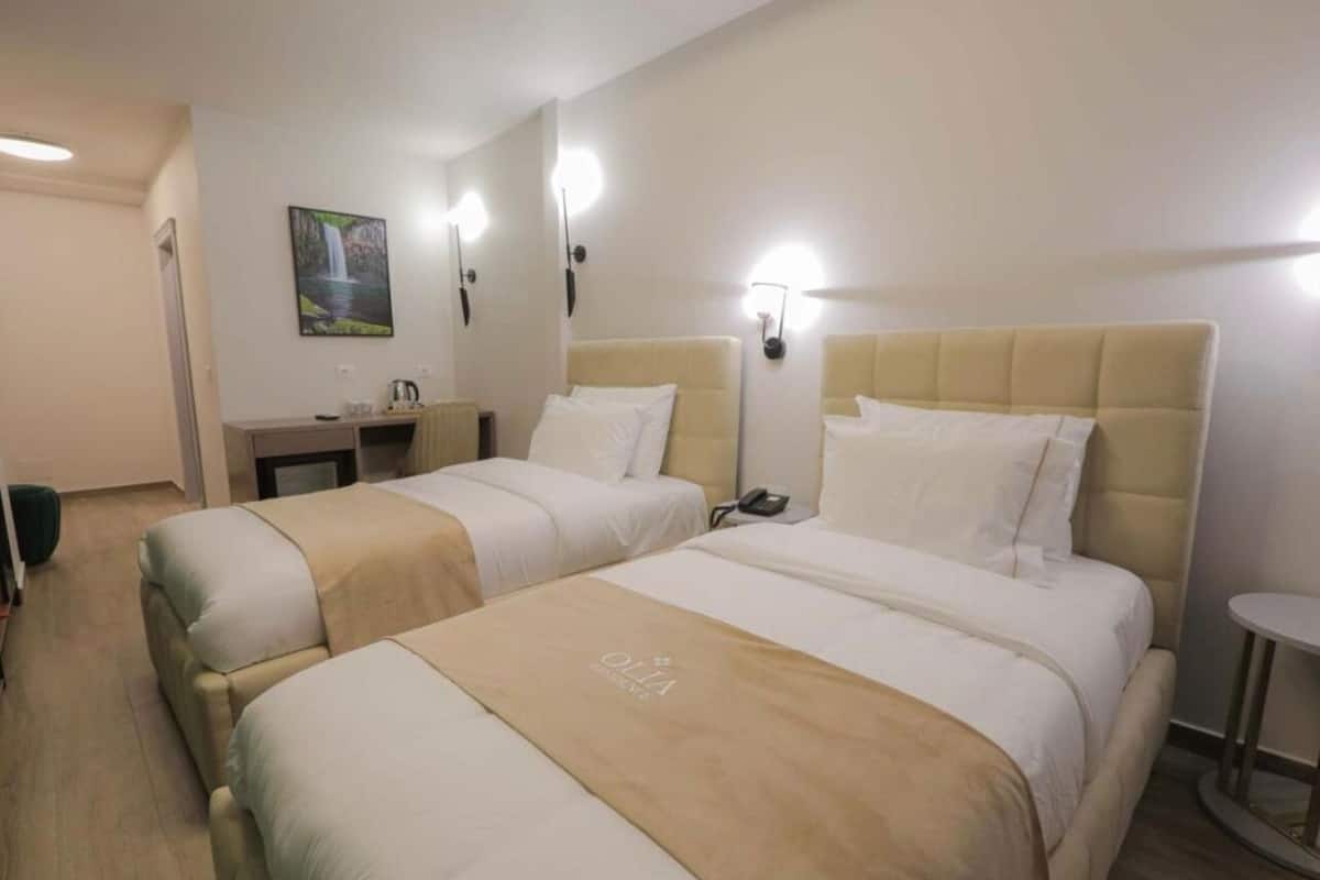 A hotel room with two beds, a small desk, a chair, and a wall-mounted light fixture.