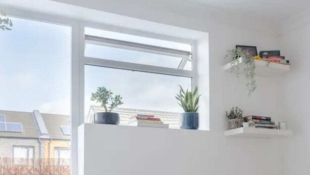 Et lyst rum med et stort vindue, hvidt møblement og hylder med bøger og planter.