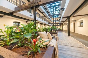 Ein großzügiger Innenhof mit hoher Decke, ausgestattet mit einem zentralen Pflanzenbeet, einer Sitzecke mit einer Couch und Kissen sowie einer Holzbank.