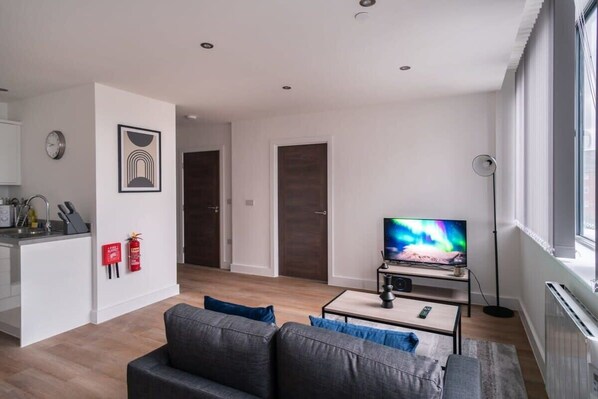 A modern living room with a grey sofa, a flat-screen TV, and a small coffee table. There is a kitchen area with white cabinets and a clock on the wall.