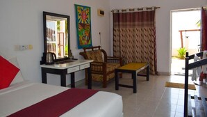 A hotel room with a bed, a desk with a chair, a small table, a colorful painting, and a window with curtains. - Golden Beach Cottages (Trincomalee)
