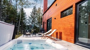 A modern wooden building with a hot tub, outdoor seating, and a forested backdrop.