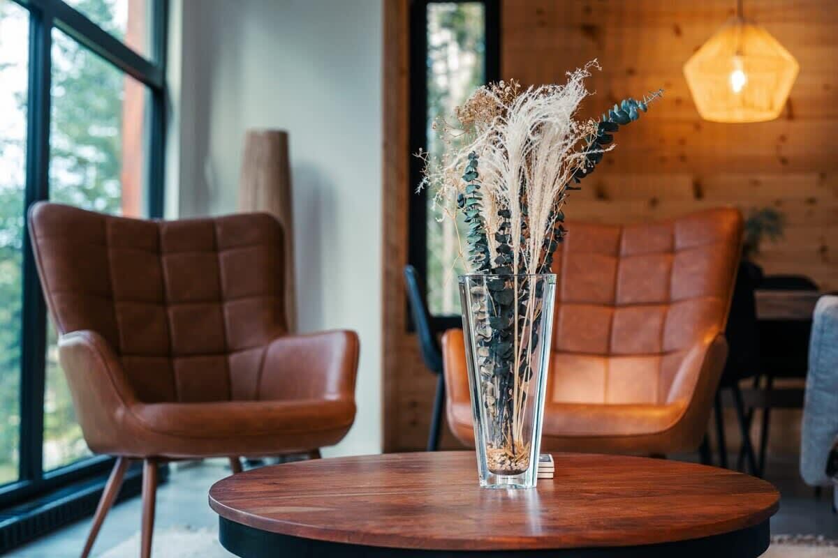 Une pièce moderne avec deux fauteuils en cuir, une table ronde en bois et un grand vase contenant des plantes séchées.