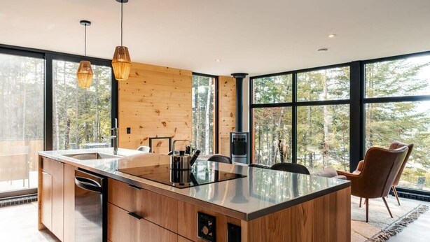 A modern kitchen with wooden cabinetry, a large island, and a view of the forest through large windows.