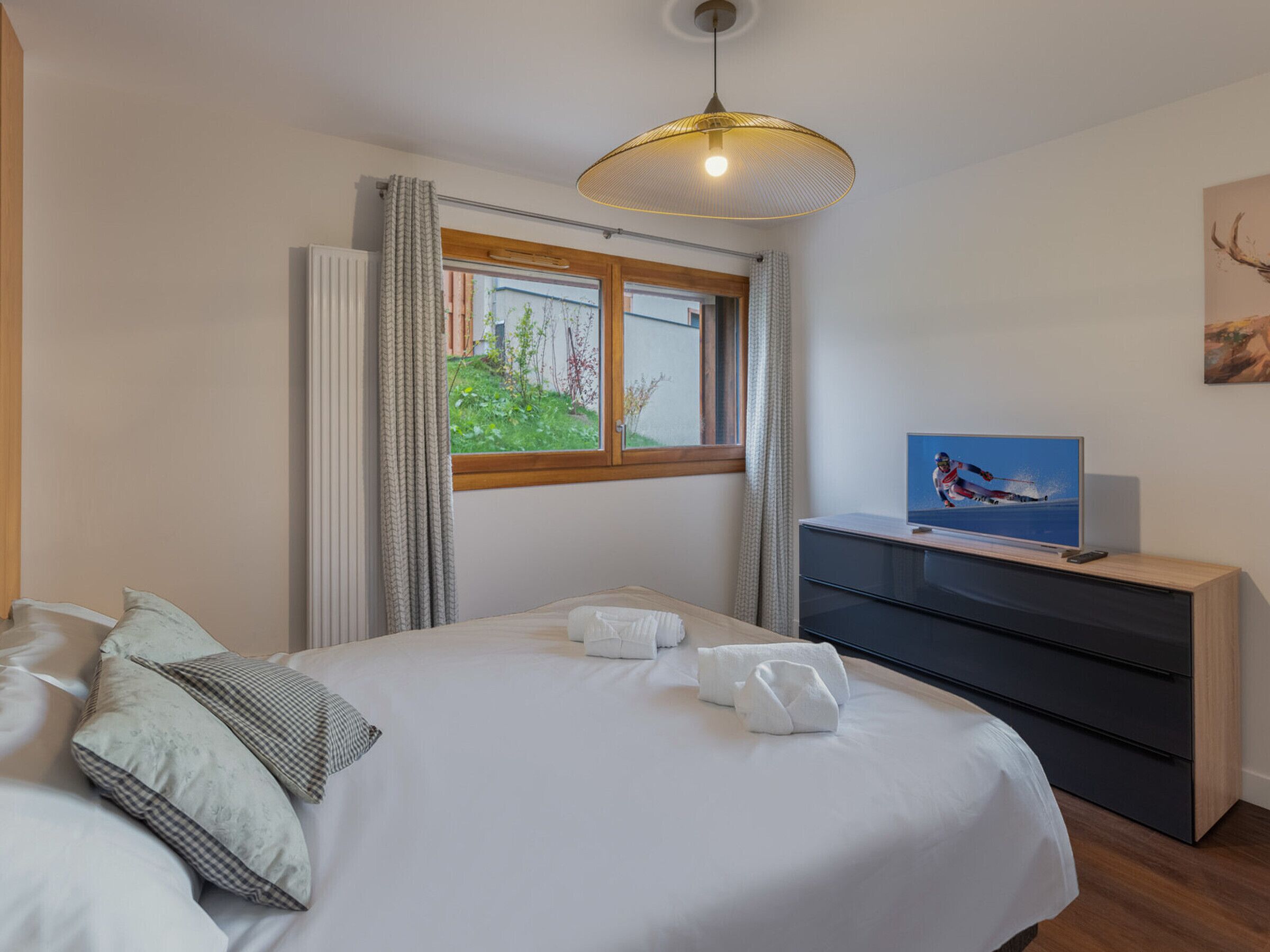 Ein Schlafzimmer mit Bett, Kommode, Fernseher und einem Fenster mit Vorhängen.