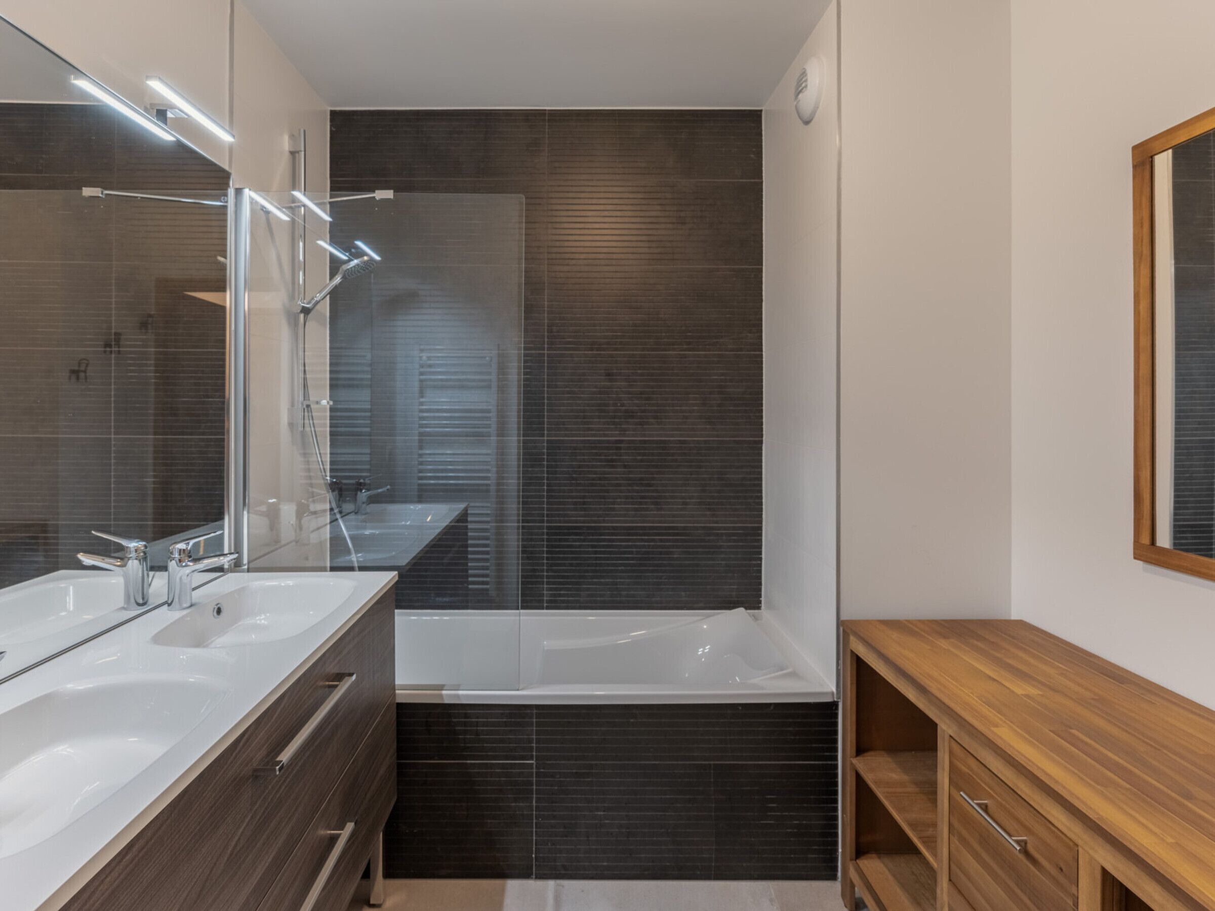 A modern bathroom with a large mirror, dual sinks, a bathtub, and a wooden vanity.
